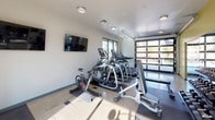 This fitness center is a thoughtfully designed, single-floor facility offering a comprehensive workout environment for a wide range of exercise needs. The space is organized into distinct functional zones, providing ample room for both cardio training and strength work without feeling cramped.

The cardio area is well-stocked with Matrix treadmills, elliptical machines, and a cycling bike, all positioned to take advantage of the abundant natural light that streams in through large sliding glass windows and a striking floor-to-ceiling glass garage door. This rollup door opens the space directly to an outdoor area, creating a seamless indoor-outdoor connection and an airy, open atmosphere.

The strength training section features a multi-station cable machine, a free weight rack with a full set of dumbbells, and adjustable benches, catering to a variety of resistance training routines. A dedicated functional fitness corner houses stability balls in multiple sizes, medicine balls, resistance bands, and yoga mats, supporting flexibility, balance, and accessory work.

Wall-mounted flat-screen televisions are positioned throughout the space for entertainment during workouts. Recessed and flush-mount ceiling lighting supplements the natural light, ensuring the facility is well-lit at all hours. The floors feature durable polished concrete with rubberized equipment mats, and the walls are accented with a bold yellow feature wall, giving the space an energetic and motivating ambiance.