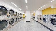 Located at 12 Marshall St in Irvington, NJ, this modern laundromat offers a generously sized, single-floor facility designed with both functionality and comfort in mind. The space is defined by its clean, contemporary aesthetic, featuring crisp white walls accented by warm golden-yellow wall panels that add a vibrant, inviting energy throughout.

The main laundry area is lined with rows of stainless steel commercial-grade front-loading washers and dryers, including stacked dryer units that maximize the use of vertical space. The machines are arranged along both walls of the elongated floor plan, creating an efficient and organized layout that accommodates a high volume of users. Yellow seating is thoughtfully positioned along one side of the space, providing comfortable waiting areas for patrons.

Underfoot, large-format light gray stone-effect tile flooring runs throughout the entire space, lending a polished and durable surface that enhances the overall cleanliness of the environment. Overhead, recessed linear LED lighting runs the length of the ceiling, casting bright, even illumination across every corner of the facility. A clean entry corridor with the same consistent finishes leads seamlessly into the main laundry hall. Structural columns punctuate the space without disrupting the open flow. The overall ambiance is bright, orderly, and modern — a well-appointed facility designed for ease of use.