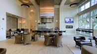 This single-floor space makes a striking impression through its soaring double-height ceilings and refined design sensibility. The open layout centers around a substantial kitchen or bar area, anchored by a bold, sculptural stainless steel range hood that extends dramatically toward the ceiling, drawing the eye upward and commanding attention as a focal point of the room. Stone-tiled wall cladding in warm beige tones frames the kitchen zone, lending a sense of permanence and luxury.

The seating area is thoughtfully arranged with woven rattan chairs and round tables, creating a relaxed yet upscale communal atmosphere. Recessed lighting and pendant fixtures punctuate the ceiling, providing layered illumination that complements the warm, neutral color palette of cream, tan, and light wood tones found throughout the space. Light hardwood-style flooring extends across the entire floor, enhancing the sense of openness and continuity.

A wall-mounted flat-screen television and a linear fireplace are integrated into the stone-clad accent wall, adding both functionality and ambiance. Circular decorative architectural elements and subtle pendant lighting contribute additional visual interest, while the overall design strikes a balance between modern elegance and comfortable, inviting warmth.
