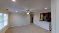 This single-floor space is designed with an open, functional layout that maximizes usability. The main living area is finished with neutral-toned carpeting and smooth walls painted in a soft, understated hue, creating a clean and versatile backdrop. Crown molding lines the perimeter of the ceiling, adding a refined architectural detail throughout. Recessed lighting is thoughtfully placed to provide even illumination, complemented by a centrally mounted ceiling fan with an integrated light fixture—ideal for both comfort and energy efficiency.

A pass-through counter connects the living area to the kitchen, offering a semi-open feel between the two spaces. The kitchen features dark wood cabinetry, a dark countertop, and stainless steel appliances including a refrigerator, contributing a modern contrast to the lighter tones of the living space. A sink is visible along the countertop near the pass-through.

Toward the back of the space, a doorway and a closet door are visible, suggesting access to additional rooms or storage. The overall ambiance is bright, tidy, and well-maintained, making it a practical and comfortable living environment.