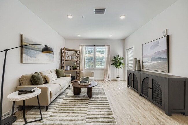 Bright and modern living room featuring a cozy beige sofa, stylish artwork, and abundant natural light from large windows. - Magnolia at Palm Bay