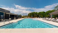 Located at 168 Headwater Circle in Mooresville, NC, this single-level community amenity space is designed for outdoor leisure and social gatherings. At its centerpiece is a large, rectangular swimming pool with crystal-clear blue water, surrounded by an expansive concrete pool deck lined with lounge chairs and shaded by blue market umbrellas — ideal for relaxing on warm Carolina days.

A generously sized covered pavilion anchors one side of the pool area, supported by sturdy brick columns with a clean, neutral-toned coffered ceiling. The pavilion provides ample shaded seating with lounge chairs and side tables, offering a comfortable retreat from the sun while maintaining views of the pool and the surrounding lush, wooded landscape.

Adjacent to the pavilion is a built-in outdoor grilling station, also framed by matching brick masonry, accompanied by open patio space and umbrella-shaded dining furniture — perfect for outdoor cookouts and community gatherings. A wrought-iron perimeter fence encloses the entire amenity area, providing security while preserving open sightlines to the natural greenery beyond. The clubhouse building, finished in a classic navy and white exterior, serves as a functional backdrop to the pool area, completing this well-appointed outdoor retreat.