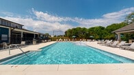 Located at 168 Headwater Circle in Mooresville, NC, this single-level community amenity space is designed for outdoor leisure and social gatherings. At its centerpiece is a large, rectangular swimming pool with crystal-clear blue water, surrounded by an expansive concrete pool deck lined with lounge chairs and shaded by blue market umbrellas — ideal for relaxing on warm Carolina days.

A generously sized covered pavilion anchors one side of the pool area, supported by sturdy brick columns with a clean, neutral-toned coffered ceiling. The pavilion provides ample shaded seating with lounge chairs and side tables, offering a comfortable retreat from the sun while maintaining views of the pool and the surrounding lush, wooded landscape.

Adjacent to the pavilion is a built-in outdoor grilling station, also framed by matching brick masonry, accompanied by open patio space and umbrella-shaded dining furniture — perfect for outdoor cookouts and community gatherings. A wrought-iron perimeter fence encloses the entire amenity area, providing security while preserving open sightlines to the natural greenery beyond. The clubhouse building, finished in a classic navy and white exterior, serves as a functional backdrop to the pool area, completing this well-appointed outdoor retreat.