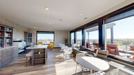 Located at 1001 Belknap St in Superior, Wisconsin, this elevated single-floor space combines a thoughtfully designed interior lounge with an expansive rooftop patio, offering panoramic views of the surrounding cityscape and beyond. The interior is defined by large, dark-framed floor-to-ceiling windows that flood the space with natural light and frame the outdoor scenery from nearly every angle. A continuous built-in window bench with storage below runs along the window wall, providing a comfortable perch to take in the views. Wood-look plank flooring adds warmth throughout, complementing the clean, neutral wall tones and recessed lighting above. One area of the interior is configured as a dedicated studio or office, featuring extensive built-in shelving, a full workstation setup, and a distinctive textured accent wall that lends character to the space. The connected rooftop patio extends the livable area outdoors, where a pergola structure provides partial shade over a composite deck. Cable-rail glass balcony fencing keeps the elevated views unobstructed, while Adirondack-style seating and a picnic table create inviting spaces for outdoor gathering. Together, the interior and exterior areas form a versatile, light-filled environment ideal for work, relaxation, or entertaining.