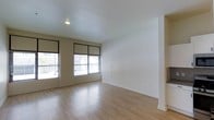 This single-floor space offers a clean, minimalist aesthetic defined by its light hardwood flooring and crisp white walls. The main living area is open and airy, benefiting from multiple windows fitted with sleek black-framed roller shades that allow for adjustable natural light throughout the day. The smooth, light-toned flooring reflects incoming daylight, enhancing the sense of openness within the room. Recessed ceiling lighting provides additional illumination, complementing the natural light and ensuring the space remains bright at all hours. The neutral color palette of the walls and flooring creates a versatile backdrop well-suited for a variety of interior styles and furnishing choices. White baseboards provide a clean border between the walls and flooring, contributing to the space's polished, contemporary feel. The layout appears straightforward and functional, with open wall space offering flexibility for furniture arrangement. Overall, the space presents a modern, well-lit environment with a calm and understated ambiance.