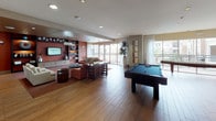 This vibrant clubroom is designed to offer a dynamic and welcoming community space across a single open floor. The layout is thoughtfully divided into distinct zones that flow seamlessly into one another. The main lounge area features warm hardwood-style flooring, plush seating arrangements with coral-toned sofas and accent chairs, a built-in entertainment wall with a large flat-screen TV, and decorative shelving with curated décor. Recessed lighting and a mix of pendant fixtures create an inviting ambiance throughout.

The game area is a standout feature, showcasing a full-size pool table and a shuffleboard table positioned near expansive floor-to-ceiling windows. Sheer white curtains frame the glass walls, allowing abundant natural light to flood the space while offering views of the surrounding outdoor area and palm trees beyond the balcony railing.

A fully equipped kitchen anchors the functional side of the clubroom, outfitted with stainless steel appliances including an oven, refrigerator, dishwasher, and coffee machines. The kitchen features eye-catching teal and copper mosaic tile backsplash, granite countertops, and warm wood cabinetry. A casual dining area with bistro-style seating provides an additional gathering spot. Together, these elements make the clubroom an ideal venue for both relaxed socializing and organized events.