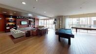 This vibrant clubroom is designed to offer a dynamic and welcoming community space across a single open floor. The layout is thoughtfully divided into distinct zones that flow seamlessly into one another. The main lounge area features warm hardwood-style flooring, plush seating arrangements with coral-toned sofas and accent chairs, a built-in entertainment wall with a large flat-screen TV, and decorative shelving with curated décor. Recessed lighting and a mix of pendant fixtures create an inviting ambiance throughout.

The game area is a standout feature, showcasing a full-size pool table and a shuffleboard table positioned near expansive floor-to-ceiling windows. Sheer white curtains frame the glass walls, allowing abundant natural light to flood the space while offering views of the surrounding outdoor area and palm trees beyond the balcony railing.

A fully equipped kitchen anchors the functional side of the clubroom, outfitted with stainless steel appliances including an oven, refrigerator, dishwasher, and coffee machines. The kitchen features eye-catching teal and copper mosaic tile backsplash, granite countertops, and warm wood cabinetry. A casual dining area with bistro-style seating provides an additional gathering spot. Together, these elements make the clubroom an ideal venue for both relaxed socializing and organized events.