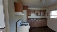 This single-story space centers around a practical kitchen that offers generous storage and a straightforward layout. Wooden cabinetry lines both the upper and lower sections of the kitchen walls, providing substantial storage capacity. The upper cabinets extend toward the ceiling, maximizing vertical space, while the lower cabinets run along the full length of the counter. Dark-toned countertops contrast with the warm wood tones of the cabinetry, and a double-basin sink is positioned along the back wall beneath the upper cabinets. A white freestanding gas range with four burners serves as the primary cooking appliance, complemented by an overhead range hood for ventilation. A louvered bi-fold door, likely concealing a pantry or utility closet, is situated adjacent to the range. The flooring throughout is a dark hardwood-style material that adds warmth to the overall aesthetic. Flush-mount ceiling lighting provides overhead illumination. The kitchen's layout is designed for efficiency, with workspace, storage, and appliances thoughtfully arranged to support everyday use.