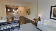 This single-floor residence offers a thoughtfully designed open-concept layout that flows effortlessly from the living area through the dining space and into the kitchen. Rich dark hardwood flooring anchors the main living areas, complemented by plush area rugs that define distinct zones within the open plan. Crown molding adds a refined architectural detail throughout, lending an air of elegance to the space.

The living area is bright and inviting, featuring neutral walls that provide a versatile backdrop for art and decor. A large colorful canvas painting adds a vibrant focal point, while recessed and pendant lighting ensures warm, layered illumination throughout. The space transitions smoothly into a dedicated dining area, which benefits from the same rich flooring and open sightlines.

The kitchen is well-equipped with stainless steel appliances, dark cabinetry, and a functional layout that includes a sink and ample counter space. Pendant lights hang above, adding both style and task lighting to the culinary space. A welcoming entryway with a clean white door completes the unit, offering a defined sense of arrival. Overall, the space conveys a modern yet warm ambiance, ideal for comfortable everyday living.