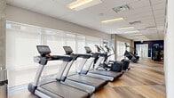 This contemporary fitness center is thoughtfully designed to accommodate a full spectrum of workout needs across multiple distinct zones. The cardio area features a row of state-of-the-art treadmills, ellipticals, and stair climbers equipped with individual digital screens, all positioned along floor-to-ceiling windows fitted with roller shades that allow for generous natural light. Light-toned hardwood-style flooring runs throughout, complementing the clean, drop-ceiling panels with recessed and flush-mounted lighting overhead. The strength training area is anchored by a bold navy blue accent wall adorned with a dynamic chevron graphic pattern, providing a visually energizing backdrop for a comprehensive selection of resistance machines, a power rack with bench, a dumbbell rack, and functional cable systems. Exposed concrete columns add an industrial character to the space. A separate functional training room, enclosed by full-height glass partition walls, houses TRX suspension trainers, medicine balls, and additional accessories. An adjoining studio area features soft sage-colored walls, open wooden shelving stocked with yoga props, and smooth floors ideal for stretching or mat-based workouts. Full-length mirrors throughout the facility enhance the sense of openness and support proper form during training.