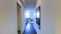 This single-floor residence showcases a sleek, modern aesthetic defined by crisp white walls, dark hardwood flooring, and an abundance of natural light. A well-proportioned hallway serves as the central artery of the home, connecting the various living areas with ease. The corridor is thoughtfully accented with a large dark-framed mirror, adding depth and visual interest while enhancing the sense of space.

The hallway leads directly into a bright and inviting living area, where floor-to-ceiling windows flood the room with natural light and offer open views beyond. The living space is furnished with a contemporary blue sectional sofa and complementary seating, creating a relaxed yet stylish atmosphere. A neutral area rug anchors the seating arrangement on the rich dark hardwood floors.

Off the hallway, a bathroom features textured wall finishes and clean, functional design elements. The overall ambiance of the residence is one of understated elegance — a harmonious blend of modern architecture and thoughtful interior design. The consistent use of a neutral white palette throughout the common areas, paired with dark flooring, creates a timeless contrast that feels both sophisticated and welcoming.