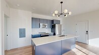 This contemporary single-floor residence showcases a thoughtfully designed open layout with a clean, modern aesthetic throughout. The kitchen is a standout feature, finished with striking slate-blue cabinetry, a white textured tile backsplash, and a full suite of stainless steel appliances including a range, over-the-range microwave, and refrigerator. A generously sized island with a crisp white quartz countertop and an undermount sink serves as both a functional workspace and a visual centerpiece, illuminated by a sculptural black sputnik chandelier with globe bulbs that adds a mid-century modern touch. Light-toned hardwood flooring runs consistently throughout the unit, enhancing the sense of continuity and warmth. The living areas benefit from large, multi-pane windows that invite generous natural light and offer views of the surrounding neighborhood. Bedrooms are bright and airy, with some rooms featuring corner windows that maximize light exposure and outdoor sightlines. Sliding closet doors provide practical storage. The bathroom is finished with warm hexagonal tile flooring, dark cabinetry with gold hardware, a white vanity countertop, and a full tub-shower combination with classic subway tile surround. The overall design balances bold color choices with a clean, minimalist palette for a polished, contemporary feel.