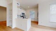 This single-floor residence presents a clean, contemporary design characterized by crisp white walls, light hardwood flooring, and an abundance of natural light throughout. The open-concept kitchen is outfitted with white shaker-style cabinetry featuring matte black hardware, a marble-style tile backsplash, white quartz countertops, and a full suite of stainless steel appliances including a range, over-the-range microwave, and refrigerator. A peninsula with an undermount sink adds functional workspace and casual dining potential. The living area is generously proportioned, with large windows and a glass-panel exterior door that invite natural light and views of surrounding greenery. The primary bedroom connects to a well-appointed en-suite bathroom featuring a dual-sink vanity with white quartz countertops, matte black fixtures, frameless glass shower enclosure, and direct access to a walk-in closet. Additional bedrooms are comfortably sized, each with a window and hardwood flooring. A dedicated laundry room with a full-size front-loading washer and dryer adds everyday convenience. Recessed lighting and ceiling fans complete the thoughtfully designed interior.