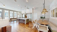 Located at 8251 Windsor Hill Blvd in North Charleston, SC, this single-floor amenity space delivers a thoughtfully designed open-concept layout that seamlessly blends function and coastal-inspired style. Rich herringbone-patterned hardwood flooring flows throughout, anchoring the space with warmth and visual depth.

The gourmet kitchen serves as a central hub, featuring a generous marble-top island with seating for multiple guests, sage-toned cabinetry, a professional stainless steel refrigerator, subway tile backsplash, and an industrial-style faucet. Overhead, mercury glass pendant lights with brass fittings add a refined touch. Large windows flood the kitchen area with natural light and provide views of the outdoor surroundings.

Adjacent to the kitchen, the dining area offers an inviting oval wood table surrounded by cane-back chairs, complemented by a built-in upholstered banquette set against a whitewashed exposed brick accent wall. Striking gold bell-shaped pendant lights and a wall mirror enhance the warmth of this gathering space.

The lounge area features high ceilings with vertical shiplap paneling, a wall-mounted flat-screen display, comfortable seating, and a built-in refreshment station. A glass-fronted market cooler adds convenient amenity access. Together, these zones create a cohesive, welcoming environment ideal for socializing and community gatherings.