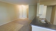 This single-floor space showcases an open-concept design that seamlessly connects the kitchen and living area. The kitchen is outfitted with white cabinetry, a dark granite-style countertop, and a stainless steel sink set into a peninsula that overlooks the main living space — an ideal configuration for both everyday functionality and casual entertaining. The living area is finished with plush wall-to-wall carpeting in a neutral tone, creating a warm and comfortable atmosphere. Crown molding runs along the ceiling throughout, adding a refined architectural detail that elevates the overall aesthetic. The walls are painted in a soft, neutral beige, contributing to a bright and welcoming ambiance. Near the entryway, two white paneled doors are visible — one serving as the front entry and the other likely a closet — providing practical storage solutions. The layout offers a natural flow between spaces, making the unit feel open and functional. The consistent use of neutral tones in flooring, walls, and trim creates a cohesive, move-in-ready environment adaptable to a variety of interior styles.