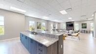 This generously proportioned single-floor communal space offers a thoughtfully designed open-plan layout that seamlessly blends multiple functional zones. The interior is finished with light wood flooring, neutral gray walls, and a suspended tile ceiling with recessed panel lighting, creating a bright and professional ambiance throughout.

At the heart of the space sits a well-appointed kitchen area, anchored by a large granite-topped island with bar seating. Dark wood cabinetry lines the back wall, complemented by stainless steel appliances including a full-size refrigerator, microwave, and ample counter space — making it ideal for casual dining or catered events.

The lounge area features a central sectional sofa arrangement around a solid wood coffee table, surrounded by a curated mix of accent chairs in warm tones. A wall-mounted flat-screen TV is set against a textured wood slat feature wall, adding warmth and visual depth to the seating zone.

A dedicated soft seating nook showcases leather armchairs gathered around a round side table, set against a striking panoramic cityscape mural, lending an urban, cosmopolitan feel. Additional high-top café-style tables and chairs are arranged near the perimeter windows, which provide abundant natural light and views of surrounding greenery. The overall atmosphere is polished, welcoming, and versatile.