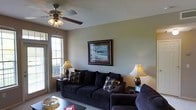 This single-floor space offers a comfortable and inviting atmosphere, highlighted by a well-appointed living room that serves as the heart of the home. The living room features neutral, earthy wall tones that create a warm and relaxed ambiance. A ceiling fan with a light fixture anchors the room from above, providing both function and a subtle decorative touch. Large windows along one wall allow natural light to flow generously into the space, brightening the interior and offering views of the surrounding exterior. The seating area is centered around a plush, dark sofa set accented with patterned throw pillows, complemented by matching table lamps that cast a soft, warm glow throughout the room. A framed coastal landscape painting displayed prominently on the main wall adds a classic, refined aesthetic to the space. The coffee table and side tables provide practical surfaces, completing the cohesive and thoughtfully arranged layout. The overall design balances comfort and style, making the living area an ideal space for relaxing or entertaining.