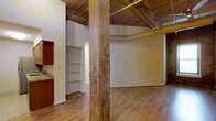 This single-floor space showcases a compelling industrial-loft aesthetic rooted in its original architectural bones. Exposed wooden ceiling planks run overhead, supported by a visible timber beam framework that lends warmth and authenticity to the interior. Thick, rustic load-bearing wood columns anchor the open layout, serving as striking structural focal points throughout the space.

The flooring is finished in smooth hardwood, providing a clean contrast to the raw, textured surfaces above. A notable exposed brick wall surrounds a window, adding further historic character and allowing natural light to filter into the interior. A ceiling fan is mounted centrally, contributing to air circulation within the open volume.

The layout includes a built-in shelving unit, offering practical storage integrated directly into the wall. A glimpse of cabinetry with granite or stone countertops hints at a kitchen or utility area nearby. An exposed HVAC duct runs along the ceiling, consistent with the space's industrial design language, where mechanical systems are left visible rather than concealed.

Overall, the space carries a warm yet raw ambiance — characteristic of a thoughtfully preserved historic building conversion — balancing rustic materials with a functional, open interior.