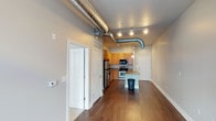 This one-bedroom, one-bathroom unit showcases a distinctive industrial aesthetic through its most eye-catching feature: large exposed silver ductwork that runs prominently across the ceiling, adding an urban loft character to the space. The open-concept layout connects the living area to the kitchen in a seamless flow, maximized by rich, dark hardwood flooring that extends throughout the main living space.

The kitchen is well-appointed with warm honey-toned cabinetry, stainless steel refrigerator, electric range, and a built-in microwave. A light-colored breakfast bar island provides additional prep space and casual dining options, acting as a natural divider between the kitchen and living area. Track lighting runs along the ceiling, complementing the industrial ductwork while providing ample illumination throughout.

The neutral gray walls create a versatile backdrop that pairs well with the warm wood tones and industrial elements. Multiple doorways are visible from the main living area, offering access to the bedroom and bathroom. The single-floor layout ensures convenient, step-free living in a compact yet functional arrangement.