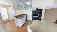 This single-floor space centers around a practical, open-concept kitchen designed for everyday functionality. The kitchen is equipped with a full suite of black appliances, including a coil-burner electric range with an overhead range hood and a side-by-side refrigerator, complemented by a black dishwasher seamlessly integrated beneath the countertop. White cabinetry lines the walls, offering generous storage both above and below the counters, while a double-basin sink sits beneath upper cabinets for added convenience. The countertops feature a speckled laminate finish in a neutral tone, extending to a peninsula or bar-style counter that helps define the kitchen space and provides additional prep or dining surface area. Warm-toned laminate wood flooring runs throughout, adding a cohesive and easy-to-maintain finish underfoot. The walls are painted in a light, neutral tone, and a textured ceiling with overhead fluorescent lighting keeps the space bright and practical. A window near the sink allows for natural light to filter in, enhancing the overall openness of the kitchen area.