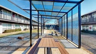 This two-story property is designed around a generously sized central courtyard that serves as the community's social hub. The buildings feature a striking exterior combination of navy board-and-batten siding and white brick, accented by sleek metal railings along the second-floor balconies. Stairways connect both levels, and individual units on the ground floor open directly onto the landscaped courtyard, while upper-floor units benefit from private balcony access and elevated views of the shared green space.

The courtyard itself is a standout feature, centered around mature deciduous trees that provide natural shade. A covered pavilion with a modern steel frame and translucent polycarbonate roof panels houses multiple picnic-style tables with wood-composite surfaces, offering a comfortable outdoor gathering space regardless of weather. An outdoor grill station and trash receptacles are conveniently positioned nearby. A fenced swimming pool area is situated at one end of the courtyard, adding a recreational amenity to the community.

Curving concrete walkways connect the various areas of the courtyard, guiding residents between the buildings, pavilion, and pool. The overall ambiance is open, social, and well-maintained, with a contemporary aesthetic enhanced by the bold color palette of the building exteriors.