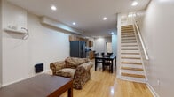 This single-floor unit is laid out in a long, efficient floor plan that connects several functional zones seamlessly. The main living area features light hardwood flooring throughout, recessed ceiling lighting, and neutral white walls that create a bright, clean atmosphere. A floral-patterned armchair and wooden coffee table anchor the living space, which benefits from a window dressed with gray curtains that admit natural light.

The open layout flows directly into a dedicated dining area, where a dark wood table and matching chairs provide ample seating. Adjacent to the dining space is a functional kitchen equipped with warm honey-toned shaker-style cabinetry, light granite-effect countertops, stainless steel appliances including a range, refrigerator, and dishwasher, plus a built-in microwave. A window above the sink adds natural light, and a rear door provides convenient exterior access.

The bedroom is generously proportioned, featuring the same hardwood flooring, recessed lighting, multiple windows with blinds, and ample room for a full bed, seating, and a work desk. A second bedroom offers a similarly open layout with a closet door and shelving. The bathroom includes a full soaking tub with floor-to-ceiling large-format tile surround and brushed metal fixtures. A staircase near the main living area suggests access to additional levels.