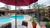 This outdoor amenity area centers around a generously sized swimming pool with clear blue water, surrounded by a wide concrete deck finished with decorative red accent strips. The pool area is enclosed by black wrought iron fencing, providing both security and an open, airy feel. Tall palm trees and mature pines frame the space, adding natural shade and a warm, resort-like atmosphere.

A substantial wood-beam pergola extends along one side of the pool deck, casting dramatic striped shadows across the ground and creating a shaded retreat for lounging or gathering. Beneath the pergola, round tables paired with slatted metal chairs offer comfortable seating, while additional chaise lounges are positioned poolside for sunbathing. Red market umbrellas are stationed throughout the deck, providing supplemental shade and adding a consistent pop of color.

The surrounding residential buildings feature tan stucco exteriors, private balconies, and exterior staircases, contributing to a cohesive community aesthetic. Manicured ground-cover plantings and potted tropical plants enhance the landscaping throughout. The overall ambiance is relaxed and inviting, balancing functional outdoor living amenities with attractive, well-tended surroundings.