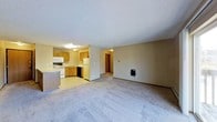 This single-floor space features an open-concept design that seamlessly connects the living area to the kitchen. The spacious carpeted living room offers a comfortable, neutral environment, with a wall-mounted utility panel adding a functional touch. A hallway visible from the main area leads to additional rooms, suggesting a well-organized floor plan.

The kitchen is outfitted with warm oak cabinetry providing ample storage, and includes essential appliances such as a white refrigerator, electric stove, built-in microwave, and a dishwasher. The countertops and sink are positioned along the cabinetry, creating an efficient workspace. The kitchen flooring transitions from the living area's carpet to a smooth, easy-to-clean surface.

Overhead lighting in both the kitchen and living space ensures consistent illumination throughout. The textured ceiling adds a subtle design element common in residential construction. The neutral wall tones throughout contribute to a versatile and functional ambiance, making the space adaptable to various interior styles.