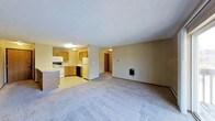 This single-floor space features an open-concept design that seamlessly connects the living area to the kitchen. The spacious carpeted living room offers a comfortable, neutral environment, with a wall-mounted utility panel adding a functional touch. A hallway visible from the main area leads to additional rooms, suggesting a well-organized floor plan.

The kitchen is outfitted with warm oak cabinetry providing ample storage, and includes essential appliances such as a white refrigerator, electric stove, built-in microwave, and a dishwasher. The countertops and sink are positioned along the cabinetry, creating an efficient workspace. The kitchen flooring transitions from the living area's carpet to a smooth, easy-to-clean surface.

Overhead lighting in both the kitchen and living space ensures consistent illumination throughout. The textured ceiling adds a subtle design element common in residential construction. The neutral wall tones throughout contribute to a versatile and functional ambiance, making the space adaptable to various interior styles.