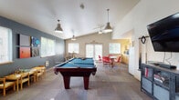 This single-floor community recreation room is thoughtfully designed to encourage social interaction and leisure. At the heart of the space sits a full-size pool table, complemented by a wall-mounted cue rack and a flat-screen television, making it an ideal hub for entertainment. Vaulted ceilings with industrial-style pendant lights and a ceiling fan lend the main area an open, airy feel, while natural light streams in through multiple windows fitted with blinds.

The space is divided into several distinct activity zones. A casual seating area features high-top bar tables with industrial-style stools, perfect for conversation or tabletop games. A separate dining nook is furnished with round tables and vibrant red chairs, adding a pop of color against the cool, dark-blue accent walls. A compact kitchenette, equipped with a refrigerator and microwave, provides convenient access to refreshments.

A cozy corner nook with a round pub-height table and bar stools offers a more intimate setting, accented by a bold dark feature wall. Patio doors provide direct access to the outdoors, further enhancing the sense of openness. The durable patterned carpet flooring ties the space together, balancing comfort and practicality throughout this well-appointed communal retreat.
