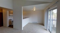 This single-floor residential space offers a clean, functional layout designed for comfortable everyday living. The living area is finished with wall-to-wall carpet in a neutral tone, creating a warm and cohesive feel throughout. A defining architectural feature is the open pass-through counter that connects the kitchen to the living area, promoting an open-concept flow while maintaining a sense of defined zones. The kitchen counter provides a convenient surface for casual dining or food preparation. Natural light enters the space through a sliding glass door or large curtained window panel, brightening the interior and offering access to the outdoors. A flush-mount ceiling light fixture provides overhead illumination in the main living area. The walls are finished in a crisp, neutral white, lending a bright and clean aesthetic that makes the space feel open and adaptable to various interior styles. A baseboard heater along the far wall ensures efficient climate control during cooler months. Overall, the space presents a straightforward and practical residential environment with a simple, understated design that offers a solid foundation for personalization.