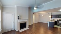 This single-story space in Frisco presents an open, flowing layout that seamlessly connects the living and dining areas. The living room serves as the focal point of the space, anchored by a classic fireplace with a white wood mantel and neutral tile surround, adding both warmth and visual interest to the room. Crown molding runs along the upper perimeter of the walls, lending an elegant, finished look throughout.

The ceilings are notably elevated, creating a generous sense of vertical space and airiness. A ceiling fan with a light fixture is centrally positioned in the living area, providing both functionality and style. Rich hardwood-style flooring extends throughout the open-plan space, offering a warm, cohesive aesthetic that ties the rooms together.

Transitioning toward the dining area, a chandelier adds a refined touch, distinguishing this zone while maintaining visual continuity with the rest of the space. The neutral, light-toned walls throughout create a clean, versatile backdrop suitable for a variety of interior styles. Overall, the space communicates a comfortable and well-appointed atmosphere with thoughtful architectural details throughout.