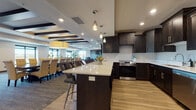 This well-appointed community amenity space is thoughtfully designed to accommodate a variety of social and functional needs. The open-concept layout seamlessly connects distinct zones, creating a cohesive and inviting environment throughout.

The full kitchen is a standout feature, outfitted with dark espresso cabinetry, white quartz countertops, stainless steel appliances including a range, dishwasher, and refrigerator, and light hardwood-style flooring. A large center island with bar seating and elegant pendant lighting serves as both a prep area and casual gathering spot.

Adjacent to the kitchen, a formal dining area accommodates a long, dark conference-style table surrounded by upholstered seating, illuminated by a statement drum pendant light. Decorative coffered ceiling details with dark wood beams add architectural character and visual depth to the space.

The lounge area offers a relaxed retreat, furnished with plush teal velvet armchairs arranged around a circular side table. Large windows flood this zone with natural light and provide views of the surrounding area, while a wall-mounted television adds functionality.

The space is completed by a well-defined entryway with dark wood doors, carpeted flooring, and clean neutral wall tones, contributing to an overall atmosphere that is both contemporary and welcoming.
