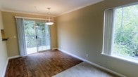 This single-floor space showcases a thoughtfully designed dining area that seamlessly connects to adjacent living spaces. The room features rich hardwood flooring that adds warmth and character, transitioning to plush carpeting in the neighboring area. Crown molding runs along the perimeter of the ceiling, lending an elegant, finished aesthetic to the space. A sliding glass door floods the room with natural light and provides direct access to an outdoor balcony, where views of mature greenery create a refreshing, private backdrop. An additional window on the adjacent wall further enhances the brightness of the interior. A pendant light fixture hangs centrally over the dining area, offering both functional illumination and a subtle decorative focal point. The walls are finished in a neutral, warm-toned paint that complements the natural wood flooring and white baseboards throughout. The overall ambiance is clean, bright, and inviting, with classic architectural details that provide a versatile foundation for a variety of interior styles.