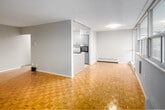 Located at 30 Speers Rd in Oakville, Ontario, this two-bedroom, one-bathroom unit offers a well-organized layout across a single floor. The entrance leads into a clean hallway with light tile flooring, providing access to the key living areas.

The living room is a standout feature, boasting a wall of large floor-to-ceiling windows that flood the space with natural light and frame views of the surrounding treetops and neighbourhood. Classic parquet wood flooring adds warmth and character, complemented by baseboard heating along the window wall.

The kitchen is thoughtfully laid out with a peninsula-style countertop featuring granite-look surfaces, a coil cooktop, stainless sink, and grey cabinetry. Tile flooring, a full-size refrigerator, and built-in oven complete the functional space.

The primary bedroom is generously sized, featuring hardwood-style flooring and a large window wall with outdoor views and baseboard heating. The second bedroom offers tile flooring, neutral walls, and a built-in closet with panelled doors, providing ample storage.

The bathroom is finished with white square wall tiles, a toilet, vanity with mirror, and chrome fixtures. Throughout the unit, the neutral colour palette and clean finishes create a bright, practical living environment.