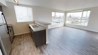 Located at 4351 15th Ave S in Seattle's vibrant 98108 corridor, this single-floor residence offers a thoughtfully designed open-concept layout that seamlessly integrates the kitchen and living areas. The kitchen features dark espresso cabinetry paired with light granite-style countertops and a double-basin sink, with a half-wall peninsula that defines the space while maintaining an open, airy connection to the living area. Durable hardwood-style flooring runs throughout, adding warmth and continuity across the entire floor plan. The living area is a standout, bathed in abundant natural light streaming through large corner windows that wrap around the room, offering expansive views of the surrounding Seattle neighborhood, including glimpses of lush tree canopies and the cityscape beyond. Horizontal blinds on each window allow for flexible light control. The clean white walls and neutral palette create a bright, modern ambiance throughout. The elevated vantage point of unit 404 enhances the sense of openness, making the space feel generous and well-connected to its urban surroundings. This residence combines practical design with contemporary finishes, offering a comfortable and inviting environment in the heart of Seattle.