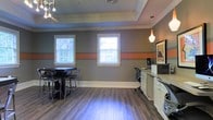 This well-appointed single-floor space combines functional design with contemporary style. The interior is defined by striking gray walls accented with a bold orange horizontal stripe, creating a modern and energetic atmosphere throughout. Crown molding and a recessed tray ceiling add architectural elegance, while warm hardwood-style flooring grounds the space with a welcoming feel.

Natural light flows in generously through multiple white-framed windows fitted with horizontal blinds, brightening the interior and offering views of the surrounding greenery. Pendant lighting with a soft, warm glow complements the recessed ceiling fixtures to create layered, versatile illumination.

The space is thoughtfully divided into distinct functional areas. A casual seating zone features a dark high-top table with matching bar stools, ideal for informal gatherings or collaborative discussions. Adjacent to this, a dedicated workstation area includes a desk, computer setup, and built-in cabinetry with ample counter space, providing a productive environment for administrative tasks. Framed artwork on the wall adds a refined, curated touch to the overall aesthetic.

The combination of stylish decor, practical furnishings, and architectural details makes this space both inviting and highly functional, suitable for a variety of uses including community meetings, work sessions, or social gatherings.