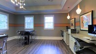 This well-appointed single-floor space combines functional design with contemporary style. The interior is defined by striking gray walls accented with a bold orange horizontal stripe, creating a modern and energetic atmosphere throughout. Crown molding and a recessed tray ceiling add architectural elegance, while warm hardwood-style flooring grounds the space with a welcoming feel.

Natural light flows in generously through multiple white-framed windows fitted with horizontal blinds, brightening the interior and offering views of the surrounding greenery. Pendant lighting with a soft, warm glow complements the recessed ceiling fixtures to create layered, versatile illumination.

The space is thoughtfully divided into distinct functional areas. A casual seating zone features a dark high-top table with matching bar stools, ideal for informal gatherings or collaborative discussions. Adjacent to this, a dedicated workstation area includes a desk, computer setup, and built-in cabinetry with ample counter space, providing a productive environment for administrative tasks. Framed artwork on the wall adds a refined, curated touch to the overall aesthetic.

The combination of stylish decor, practical furnishings, and architectural details makes this space both inviting and highly functional, suitable for a variety of uses including community meetings, work sessions, or social gatherings.