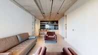Located at 2820 W Broad St in Richmond, Virginia, this single-floor lounge space combines raw industrial character with refined modern finishes. The exposed concrete ceiling, complete with visible ductwork, conduit, and track lighting, sets a striking architectural tone throughout the open layout. Polished concrete floors flow seamlessly from one end to the other, reinforcing the space's cohesive industrial aesthetic.

The main lounge area is furnished with a mix of tufted leather and velvet seating, including a full sofa, armchairs, and additional accent chairs, creating a comfortable and inviting gathering environment. A standout feature is the bold black marble-paneled wet bar at the far end, flanked by white cabinetry and a counter with bar-height seating — ideal for entertaining or informal meetings.

Large floor-to-ceiling glass windows at one end allow natural light to pour in, offering views of the surrounding West Broad Street corridor. A built-in wood shelving niche is recessed into one of the white walls, adding a warm, functional design detail. A restroom is accessible via a hallway near the entrance, with an exposed concrete column accentuating the industrial character of the corridor. The overall ambiance is sophisticated yet approachable, blending urban edge with comfortable functionality.