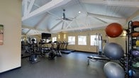 This fitness center offers a spacious, single-floor layout beneath striking vaulted ceilings with white-painted exposed beams, lending the space an open and airy feel. A ceiling fan mounted at the peak ensures comfortable air circulation throughout workouts. The flooring consists of durable rubber matting, ideal for high-traffic exercise use. Natural light streams in through a row of windows along one wall, providing views of the surrounding exterior while keeping the environment bright and inviting. Warm wall sconces complement the natural light for a well-lit atmosphere at any hour. The cardio section is well-appointed with multiple treadmills and elliptical machines, each equipped with individual display screens. A large flat-screen television is mounted on the wall, offering entertainment during workouts. A wall of mirrors along one side visually expands the space and assists users in monitoring their form. The strength training area includes a multi-function cable machine and stability balls, rounding out the available equipment. The warm yellow walls contribute to an energizing yet comfortable ambiance, making this fitness center a functional and inviting space for daily exercise.