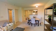 This single-floor residence offers a clean, functional layout with a warm and inviting ambiance throughout. The open-concept dining area serves as a central gathering space, furnished with a classic dark-framed dining table and elegant upholstered chairs, ideal for both casual meals and entertaining. Soft, neutral-toned carpeting extends across the living and dining areas, contributing to the overall sense of warmth and comfort.

A pass-through window connects the dining space to the kitchen, allowing natural interaction between the two areas while maintaining a sense of definition. The kitchen features white cabinetry, dark countertops, and standard appliances, providing a practical and tidy workspace. A small touch of greenery on the counter adds a fresh, lively accent to the room.

The entryway is visible from the dining area, offering a smooth and accessible transition into the main living spaces. Flush-mount ceiling lighting throughout provides consistent, even illumination across each room. The overall color palette — soft whites, creams, and beiges — creates a cohesive and calming atmosphere. The thoughtful layout and neutral finishes make this a versatile space well-suited to a variety of lifestyles and furnishing preferences.