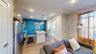 This single-floor residence is designed with a contemporary open-concept layout that seamlessly connects the kitchen, dining, and living areas. The kitchen is well-appointed with light wood cabinetry, stainless steel appliances, a white subway tile backsplash, and a granite-topped island that doubles as a breakfast bar with pendant lighting above. A bold teal accent wall adds a vibrant focal point to the dining and kitchen zone, which also features access to an exterior door.

The living area offers a comfortable, functional layout with wood-style plank flooring, recessed lighting, and a dedicated space for a media setup and workspace with open shelving. Natural light filters in through windows dressed with sheer curtains, enhancing the bright and airy ambiance.

A wide hallway connects the main living spaces to the private quarters, where the bedroom features plush carpeting, a ceiling fan, and a generously sized walk-in closet. Large windows in the bedroom allow for ample natural light.

The bathroom is finished with a wide double-sink vanity topped with granite, warm wood cabinetry, a large mirror with overhead lighting, and light tile flooring, delivering a clean and polished aesthetic throughout.