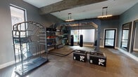 Located at 2501 Via Romana in Fort Worth, TX, this fitness center is a comprehensively outfitted single-floor facility designed to support a wide range of training disciplines. The space is divided into distinct workout zones, each thoughtfully arranged for both functionality and aesthetics.

The primary training area is anchored by a large industrial-style functional fitness rig, complete with suspended heavy punching bags, battle ropes, and TRX suspension trainers. Nearby, a fully stocked wall-mounted storage system holds medicine balls, resistance bands, and additional accessories. Plyo boxes are positioned centrally for high-intensity interval training. A stair climber unit and agility ladder rounds out this section's dynamic training offerings.

An adjacent open zone features a WaterRower rowing machine, a SCI FIT recumbent exercise bike, and free weights, with carpet tile flooring throughout providing grip and comfort underfoot. Exposed concrete beams and moody slate-blue walls lend the space a modern, industrial character, while pendant lighting fixtures cast focused illumination across the floor.

A mounted flat-screen TV and a built-in water fountain station serve as convenient amenities within the space. Floor-to-ceiling glass sliding doors connect the workout areas, allowing natural light to filter through and creating an open, airy feel. The overall ambiance is polished yet purposeful, making this fitness center a premium destination for serious training.