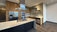 This contemporary two-story residence offers 2,230 square feet of thoughtfully designed living space, with 1,090 sq ft on the ground floor and 1,130 sq ft on the upper level. The open-concept kitchen is a standout feature, boasting warm wood cabinetry, quartz countertops with a mosaic tile backsplash, stainless steel appliances including a double wall oven, built-in microwave, gas cooktop with an overhead island range hood, and a deep double-basin sink with floating stainless steel shelves. The living room is bathed in natural light through expansive floor-to-ceiling windows and sliding glass doors that frame sweeping mountain and neighborhood views, with plush carpeting underfoot and recessed lighting above. The primary bathroom is a refined retreat featuring large-format dark tile, a soaking tub, and a private water closet, while the secondary bathroom offers a warm wood vanity with quartz countertop and a large mirror. Two well-sized closets, including a walk-in with built-in shelving and hanging rods, provide ample storage. Additional spaces include a carpeted entry area with recessed lighting, a connecting hallway, and a staircase linking both floors. Dark hardwood-style flooring flows through the main living areas, complementing the unit's modern, neutral aesthetic throughout.