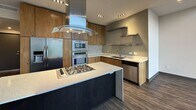 This contemporary two-story residence offers 2,230 square feet of thoughtfully designed living space, with 1,090 sq ft on the ground floor and 1,130 sq ft on the upper level. The open-concept kitchen is a standout feature, boasting warm wood cabinetry, quartz countertops with a mosaic tile backsplash, stainless steel appliances including a double wall oven, built-in microwave, gas cooktop with an overhead island range hood, and a deep double-basin sink with floating stainless steel shelves. The living room is bathed in natural light through expansive floor-to-ceiling windows and sliding glass doors that frame sweeping mountain and neighborhood views, with plush carpeting underfoot and recessed lighting above. The primary bathroom is a refined retreat featuring large-format dark tile, a soaking tub, and a private water closet, while the secondary bathroom offers a warm wood vanity with quartz countertop and a large mirror. Two well-sized closets, including a walk-in with built-in shelving and hanging rods, provide ample storage. Additional spaces include a carpeted entry area with recessed lighting, a connecting hallway, and a staircase linking both floors. Dark hardwood-style flooring flows through the main living areas, complementing the unit's modern, neutral aesthetic throughout.