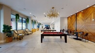 This expansive single-floor space is designed as a contemporary amenity and social hub, blending sophisticated design with versatile functionality. Large floor-to-ceiling windows line the perimeter, flooding the interior with natural light and offering views of the surrounding landscape, while hanging greenery and potted plants add organic warmth throughout.

The main lounge area is anchored by a striking sculptural gold chandelier suspended above a full-size billiards table set atop large-format, light-toned stone tile flooring. Adjacent to the game area, a dramatic chevron-patterned wood accent wall creates a bold focal point and frames a bar-height seating area with high-top tables and stools. A built-in credenza provides additional storage and surface space nearby.

A dedicated media lounge features a four-panel TV display mounted on a textured stone accent wall, complemented by a sleek blue sectional sofa and marble-topped coffee tables, ideal for group viewing or casual gatherings.

Near the entrance, a welcoming reception area includes a front desk, vibrant wall art, and a leather lounge seating arrangement. A glass-enclosed conference room with a full boardroom table offers a private setting for meetings. A kitchenette with cabinetry, a beverage station, and counter space rounds out the functional amenities of this well-appointed space.