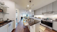 This thoughtfully designed single-floor residence offers a seamless blend of modern style and functional living. The entry foyer sets an elegant tone with rich hardwood flooring, raised-panel white doors, and a decorative console area that welcomes you into the space. The open-concept kitchen is a true centerpiece, featuring grey shaker-style cabinetry, a dramatic dark granite island with bar seating, a classic white tile backsplash, stainless steel gas range, and eye-catching pendant lighting. The kitchen flows effortlessly into a dining area and a bright, airy living room anchored by expansive windows that invite generous natural light and open outdoor views. The primary bedroom is a serene retreat with plush carpet, large windows framing outdoor scenery, and direct access to a well-appointed bathroom complete with a glass-enclosed walk-in shower finished in large-format tile, alongside a spacious walk-in closet with built-in shelving. A second bedroom offers the same soft carpet and clean, neutral aesthetic. Hardwood floors, recessed lighting, and a consistent soft white palette tie each room together, creating a cohesive and contemporary ambiance throughout.
