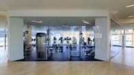 This contemporary fitness facility is designed with both functionality and aesthetics in mind, offering a welcoming and motivating environment for exercise. The standout architectural feature is the expansive floor-to-ceiling glass wall system that separates the main workout area, providing a sense of openness while offering clear sightlines into the fully equipped gym space. Natural light floods the interior through the large glass panels, which also frame views of the lush outdoor greenery beyond — creating an energizing atmosphere that blurs the boundary between indoors and out. The gym floor is fitted with professional-grade rubber flooring for safety and performance, contrasting with the warm light wood flooring in the adjacent area. The workout space is well-appointed with a comprehensive range of strength and cardio equipment, including cable machines, resistance stations, treadmills, and elliptical trainers, catering to a wide variety of fitness routines. Overhead linear lighting ensures the space is evenly and brightly illuminated throughout. The clean, minimalist design with white walls and modern finishes gives the facility a polished, professional feel, making it a well-rounded and fully functional space suited for both individual training and general fitness use.