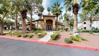 This single-story community presents a striking Spanish-inspired architectural style, characterized by warm stucco exteriors, red tile rooflines, arched entryways, and decorative detailing throughout. The welcoming clubhouse entrance is framed by tall palm trees and carefully maintained desert landscaping, including ornamental shrubs, native ground cover, and seasonal flowering plants, creating an inviting curb appeal.

The centerpiece of the outdoor amenity area is a generously sized freeform swimming pool with crystal-clear turquoise water. A standout feature is the large rock waterfall structure positioned at the heart of the pool, constructed from natural stone and designed to cascade water into the pool below, adding both visual drama and a soothing ambiance. The expansive pool deck surrounds the water on multiple sides, offering ample space for sunbathing and relaxation, complete with lounge chairs positioned to take full advantage of the open sky and abundant sunlight.

Adjacent to the main pool is a separate spa, finished with decorative stone surroundings and equipped with entry handrails. The entire outdoor area is enclosed by fencing and framed by a lush canopy of mature palm trees and evergreens, providing shade and a resort-like atmosphere throughout.