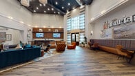 Located at 200 E Cama St in Charlotte, NC, this single-floor amenity and lounge space is a thoughtfully designed communal hub that blends industrial architecture with warm, contemporary interiors. The main lounge area commands attention with its dramatically high ceilings, exposed dark ceiling structure, and an abundance of recessed lighting that creates a welcoming ambiance. Wide-plank hardwood flooring runs throughout, grounding the space with a natural, earthy tone.

Seating is arranged in curated vignettes featuring tufted leather lounge chairs in cognac tones, plush teal sofas, and mid-century-inspired wooden accent chairs layered over area rugs. A dedicated lounge nook showcases banquette seating upholstered in rich amber velvet, accented by an eclectic gallery wall with a bold "LOUNGE" sign as its centerpiece.

The built-in bar area is equipped with warm wood cabinetry, a white quartz countertop, a black matte sink, subway tile backsplash, and wall-mounted flat-screen TVs set against a striking textured wood-panel accent wall. Floor-to-ceiling windows and a set of elegant French doors flood the entry with natural light, framed by flowing patterned drapery and flanked by lush potted plants. A chandelier-lit elevator lobby with large-format tile flooring and curated artwork completes the space.